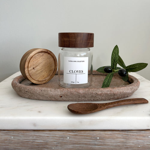 Spice Jar with wooden acacia lid  and white label and spoon on a stone tray against a neutral background