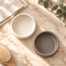 Load image into Gallery viewer, Two round ceramic trays for soap dispenser, one in warm off-white and one in textured dark gray, arranged on a beige marble surface with a folded cream towel, eucalyptus leaves, and a wooden brush nearby.
