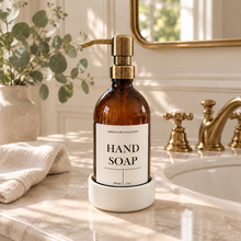 Load image into Gallery viewer, An amber glass hand soap bottle with a gold pump and a white label reading 'HAND SOAP' stands on a white marble sink base on a bathroom counter in a white ceramic  round tray A folded cream towel, a vase with greenery, and a brass faucet are visible in the softly lit background.
