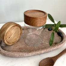 Load image into Gallery viewer, Glass jar with wooden lid on a stone tray with olive leaves and black olives.
