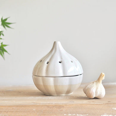White ceramic garlic canister holder on a wooden surface with a small garlic bulb and green leaves in the background.
