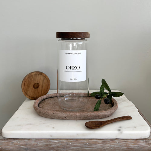 Glass jar with acacia wood lid with a white waterproof label on a marble surface with olive branch and wooden spoon.