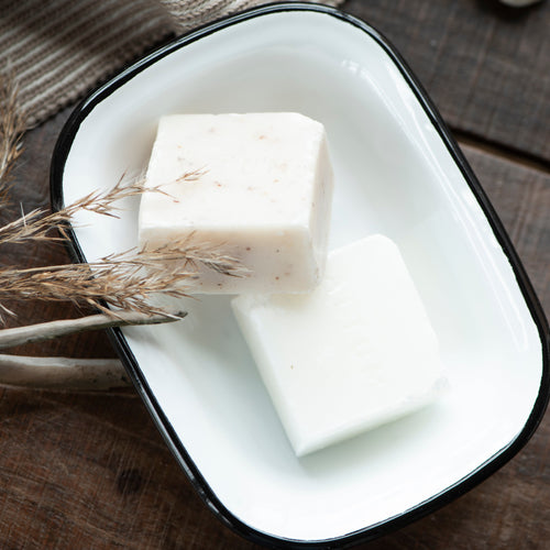 Two white bars of soap in a black-rimmed white enamel soap dish on a wooden surface with a textured fabric background.