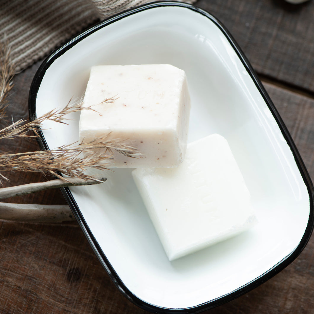 Two white bars of soap in a black-rimmed white enamel soap dish on a wooden surface with a textured fabric background.
