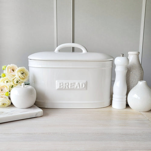 A white ceramic bread bin with 'BREAD' text embossed on the front, placed on a kitchen counter with decorative items and a vase of flowers beside it.
