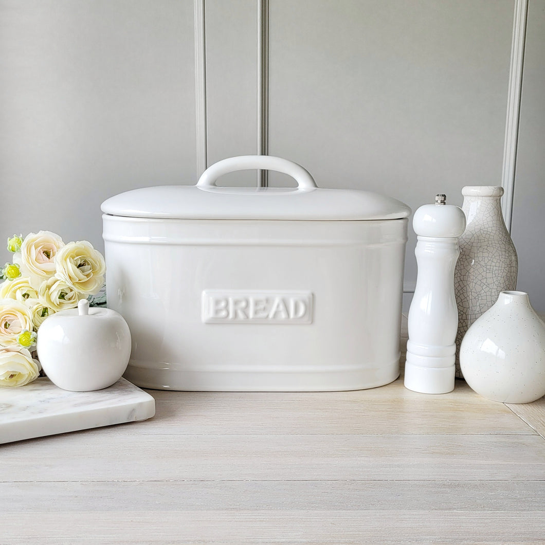 A white ceramic bread bin with 'BREAD' text embossed on the front, placed on a kitchen counter with decorative items and a vase of flowers beside it.